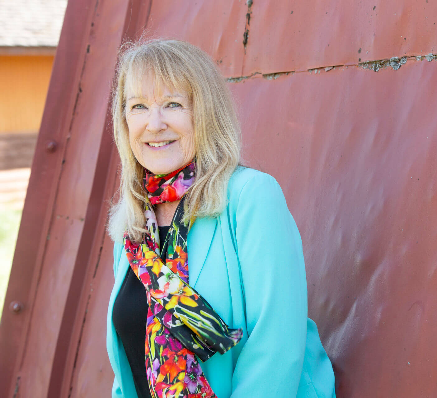 Photo of Judy Molland taken in Missoula, Montana. Judy is leaning against a red somewhat rustic building.